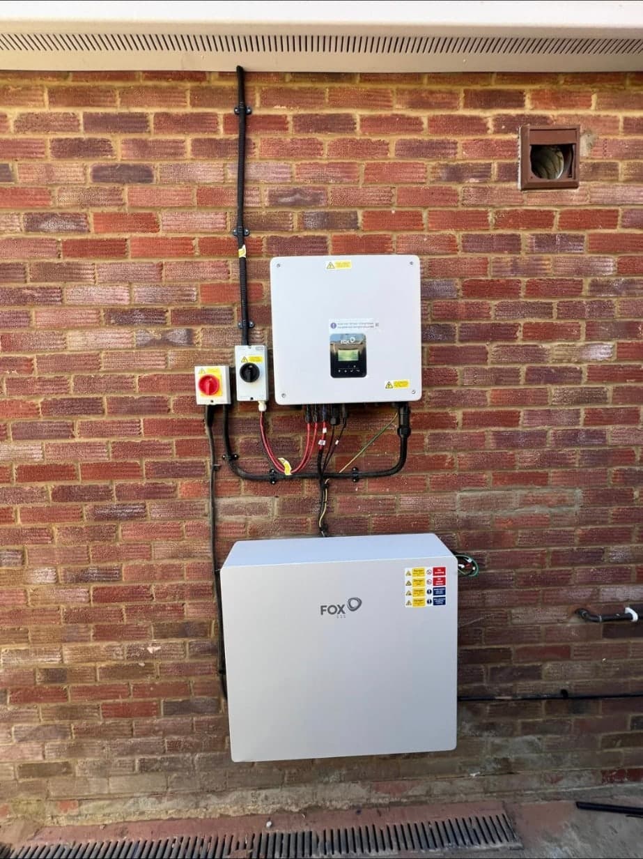 A Fox ESS home battery mounted on a brick wall beneath its hybrid inverter, with isolators and labelled DC cabling neatly clipped between them.