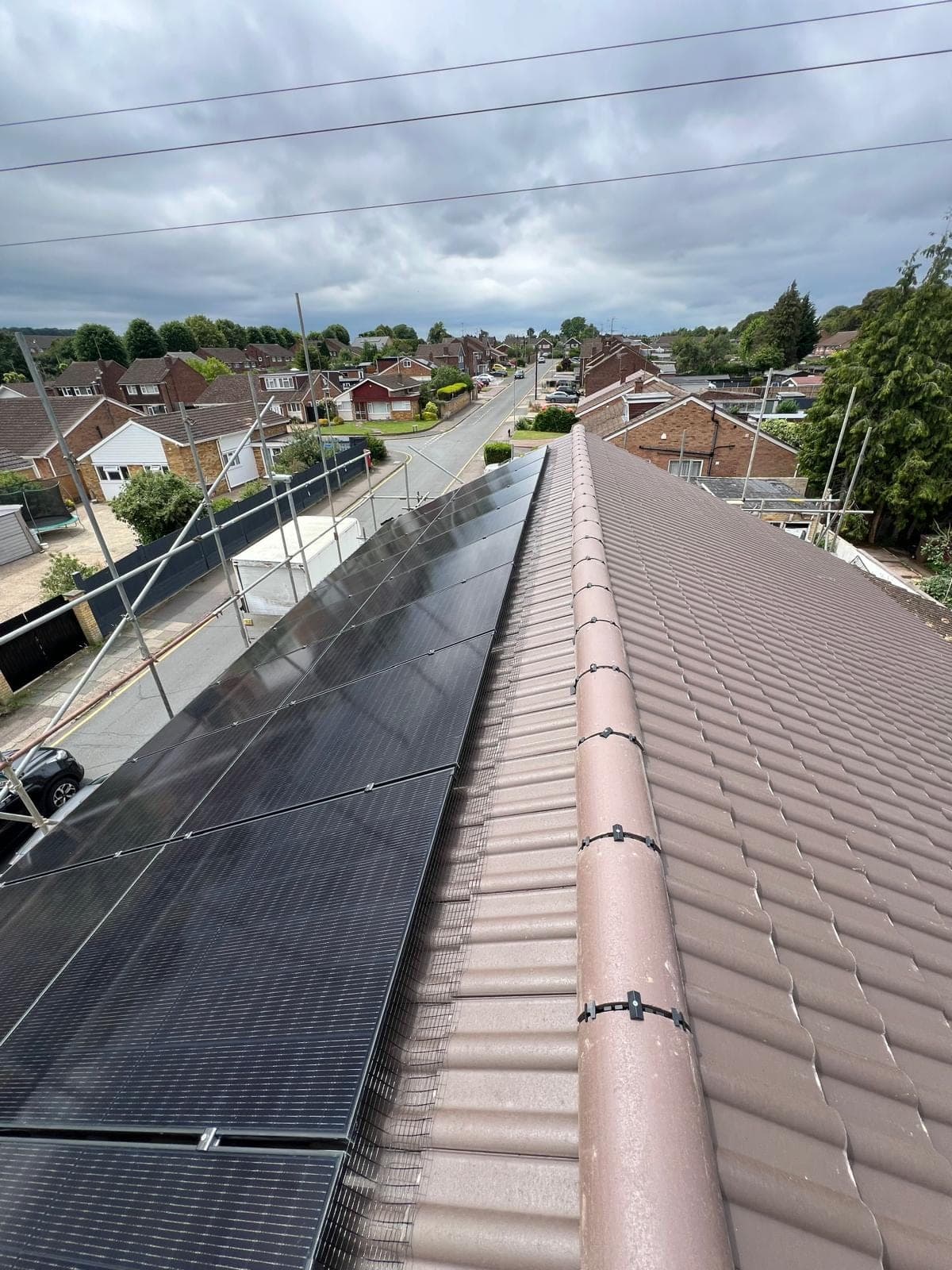 A solar photovoltaic array part-way through installation on a tiled roof, seen along the ridge, with scaffolding in place around the property and bird protection mesh fitted along the edge of the panels.